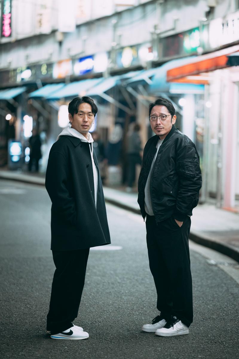 繁華街で立ち止まり対話する黒いジャケット姿の男性二人［モデル：大川竜弥 ゆうせい］
