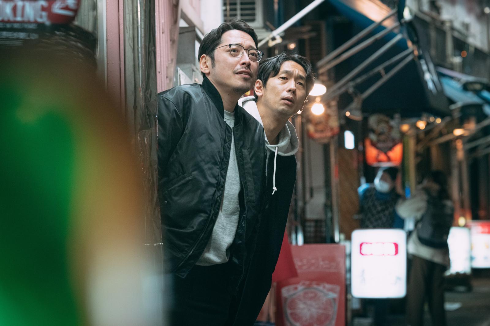 壁に身を寄せて上を見つめる眼鏡姿の男性と耳にイヤホンをつけた男性［モデル：大川竜弥 ゆうせい］