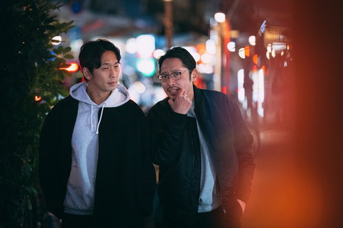 夜の繁華街で調査にあたる探偵のふたり、ネオン商店街を歩く