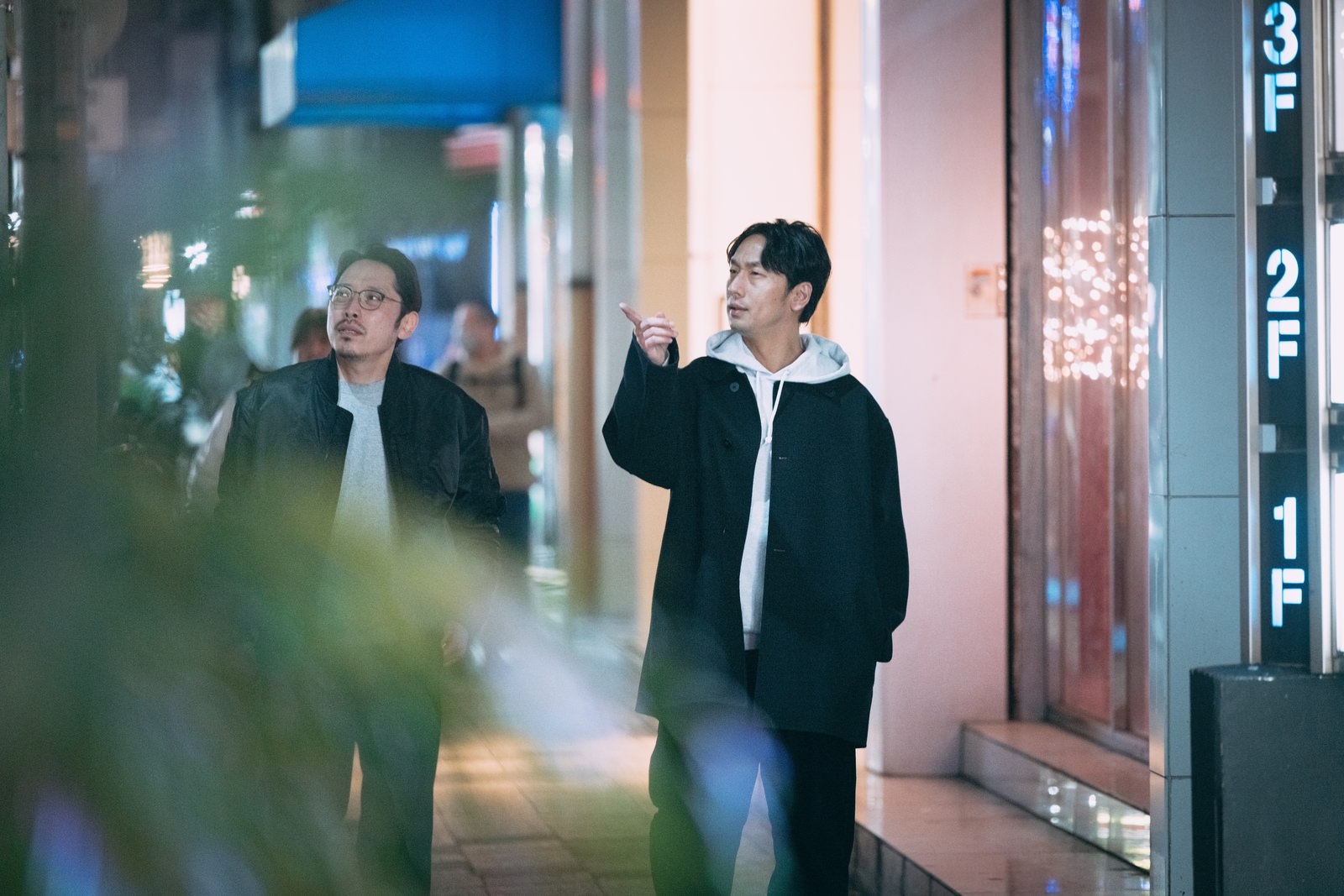 夜の繁華街で飲み屋を探す眼鏡の男性と黒いジャケットの男性［モデル：大川竜弥 ゆうせい］