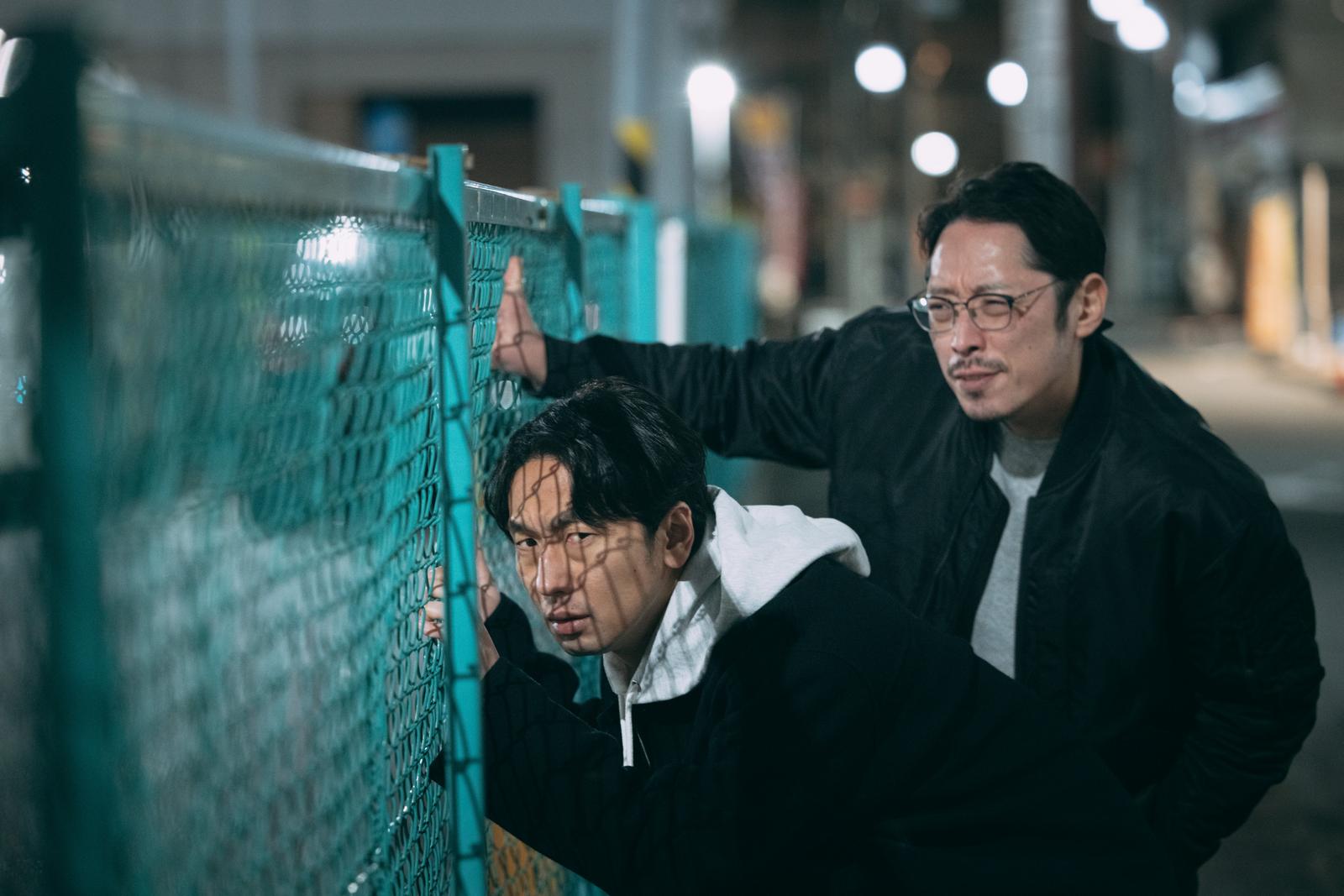 夜間にターコイズ色のフェンス越しに何かを監視する眼鏡をかけた二人の男性［モデル：大川竜弥 ゆうせい］