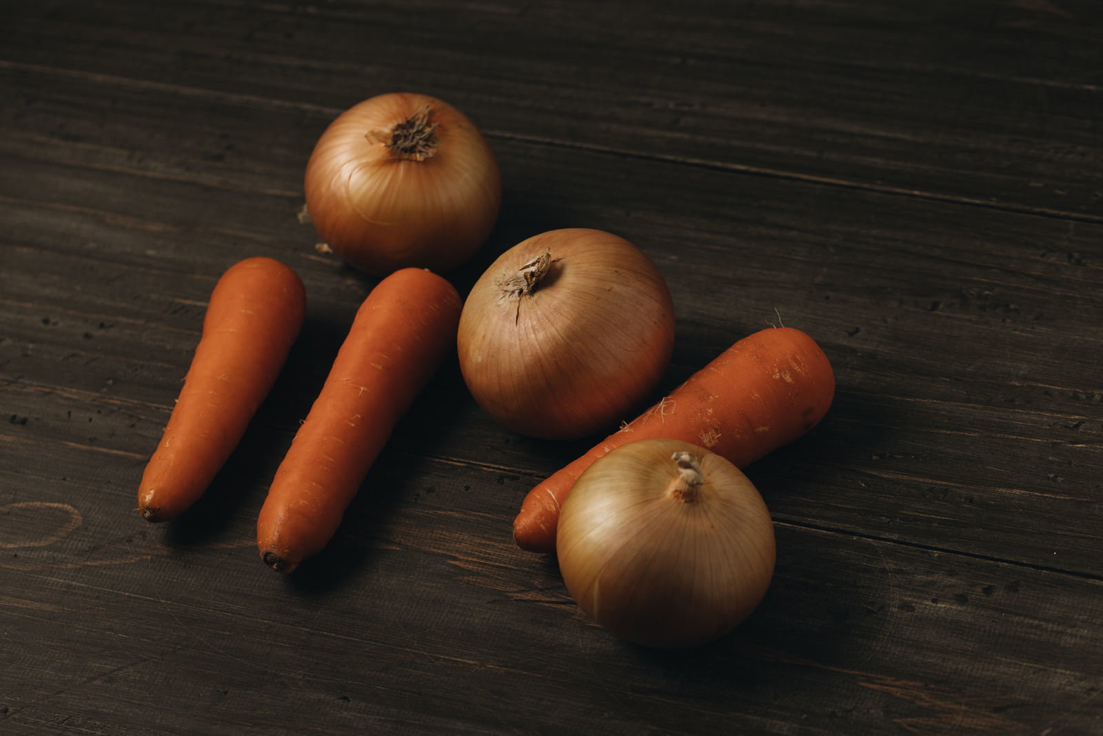 暗い木目の表面に無造作に置かれたオレンジ色の人参3本と茶色の玉葱3個
