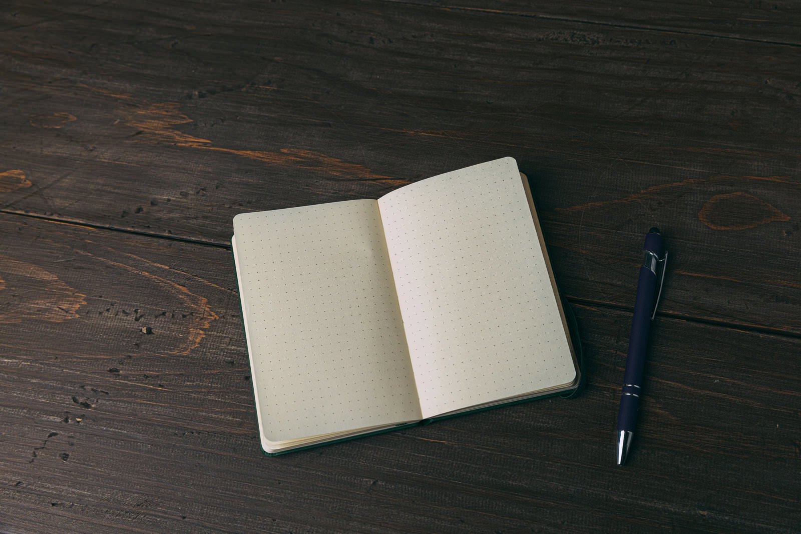 濃い木製テーブルの上に置かれた、開いた緑色カバーの手帳と黒いボールペン