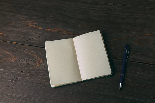 ボールペンと白紙の手帳を木製テーブルに置いた光景
