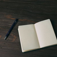 濃紺のボールペンと開いた白紙の手帳が置かれた木目調のテーブルの写真