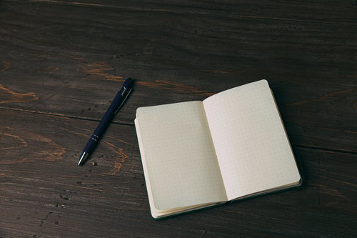 濃紺のボールペンと開いた白紙の手帳が置かれた木目調のテーブル