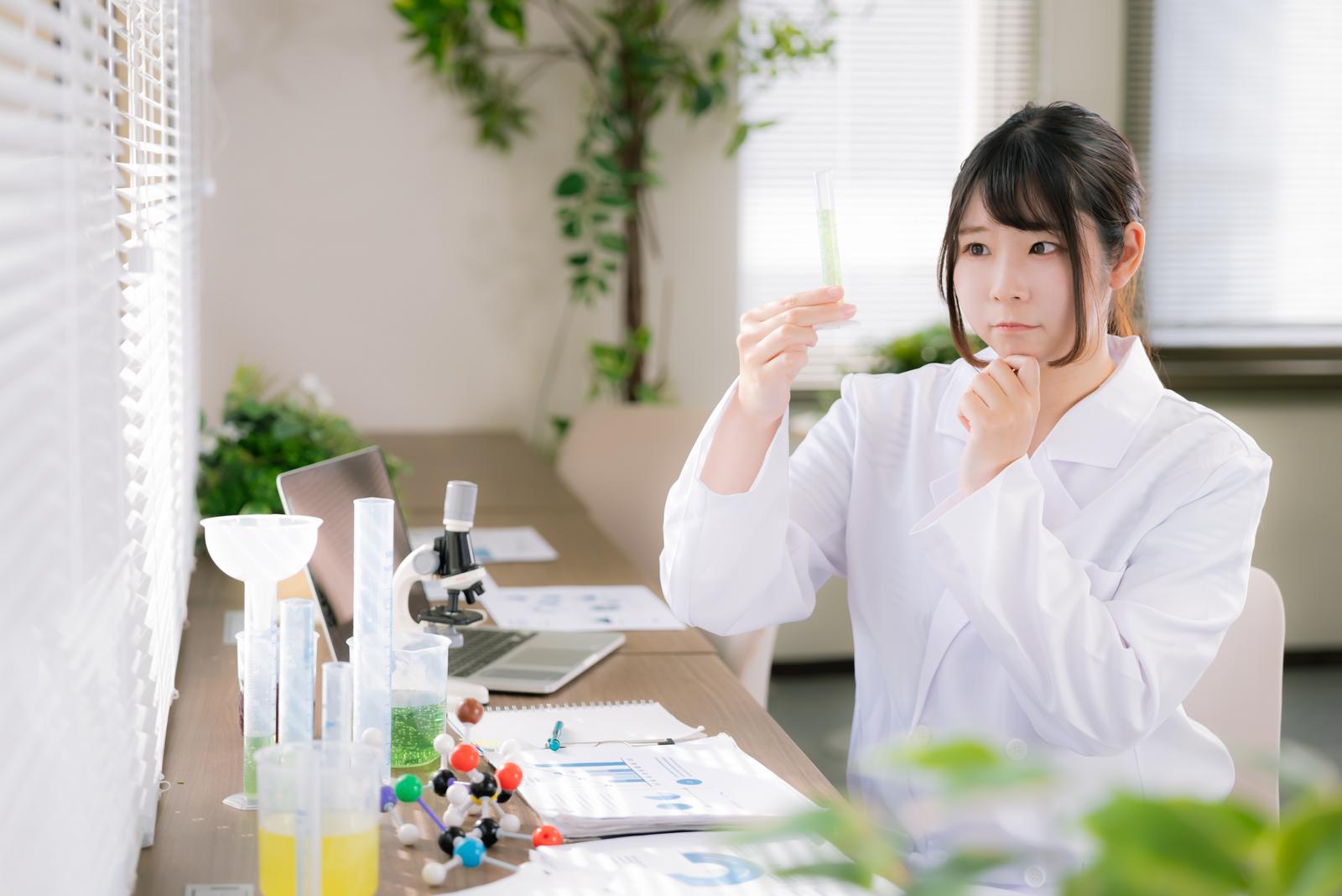 白衣を着た女性が試験管を持ち、グリーン色の液体を観察している実験室の風景［モデル：モデルリリース］