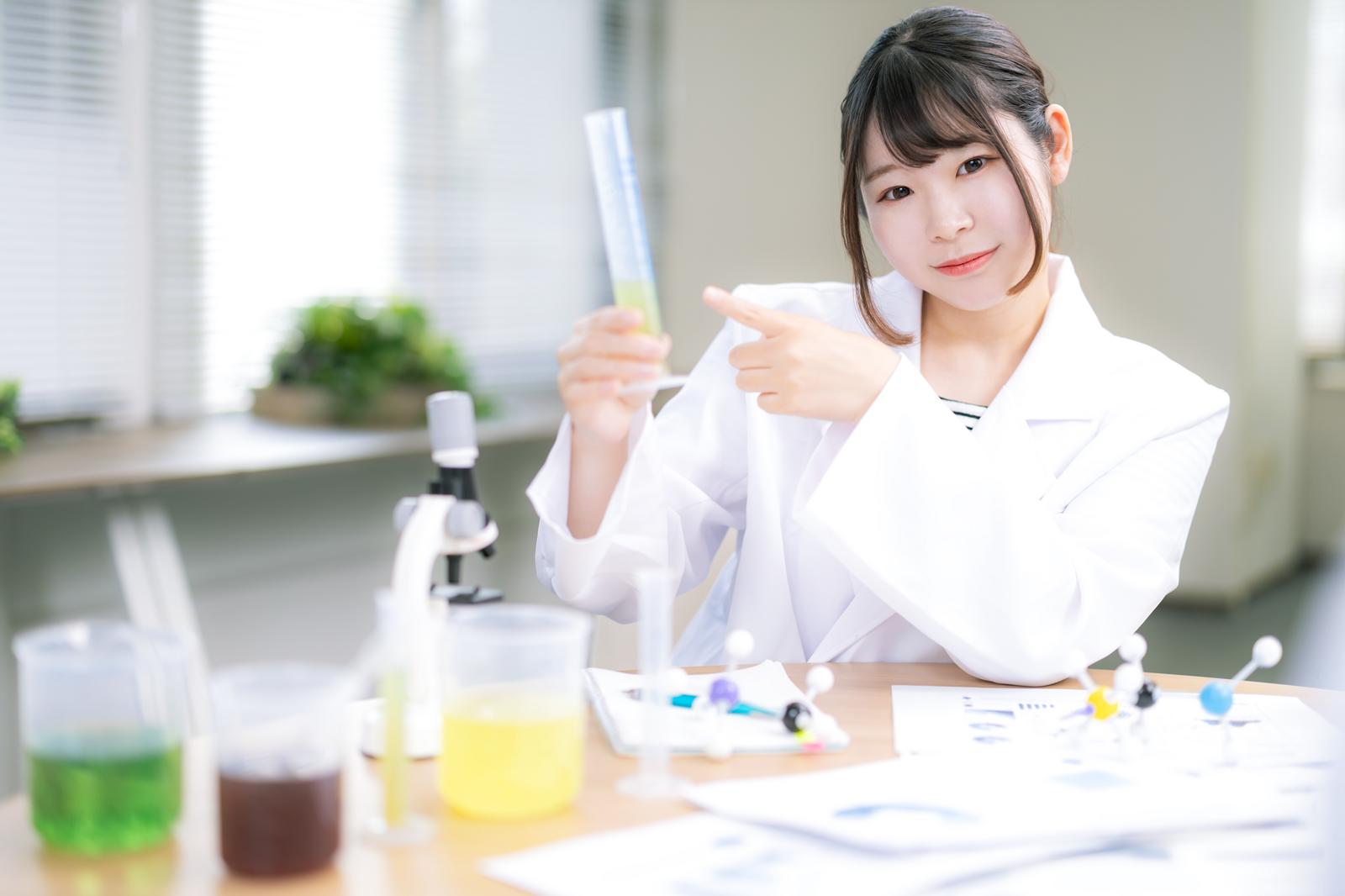 研究室で試験管を持ち顕微鏡の前に座る白衣姿の女性研究者［モデル：モデルリリース］