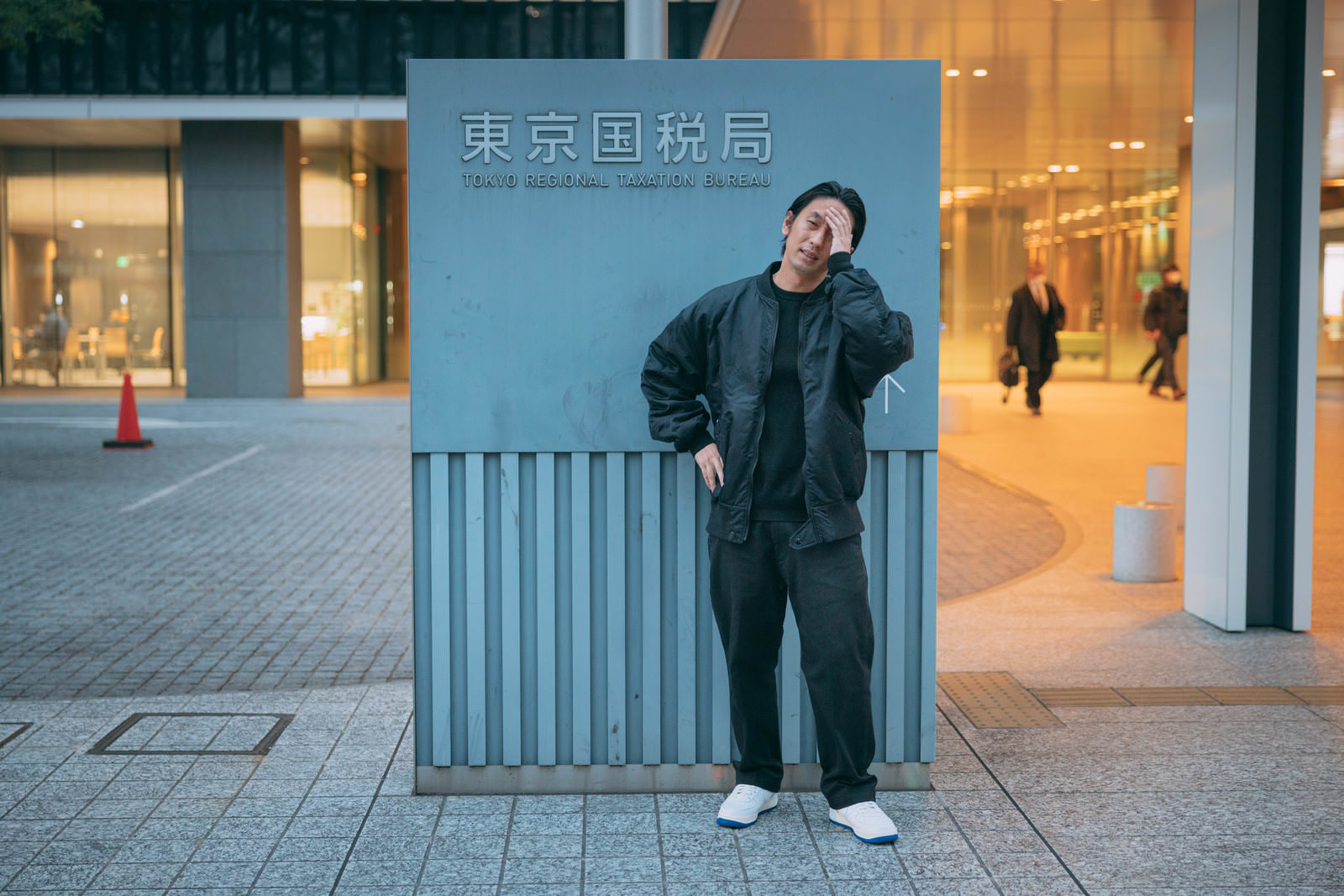 東京国税局の看板の前で頭を抱えて困る黒いコートの男性［モデル：大川竜弥］
