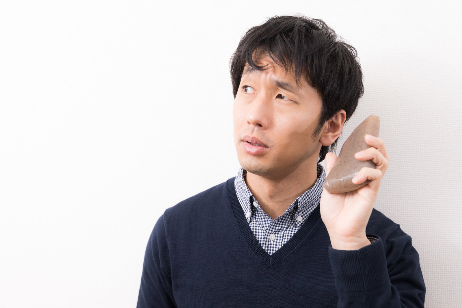 こんにゃくを耳に当てて困った表情を見せる紺色セーターの男性［モデル：大川竜弥］