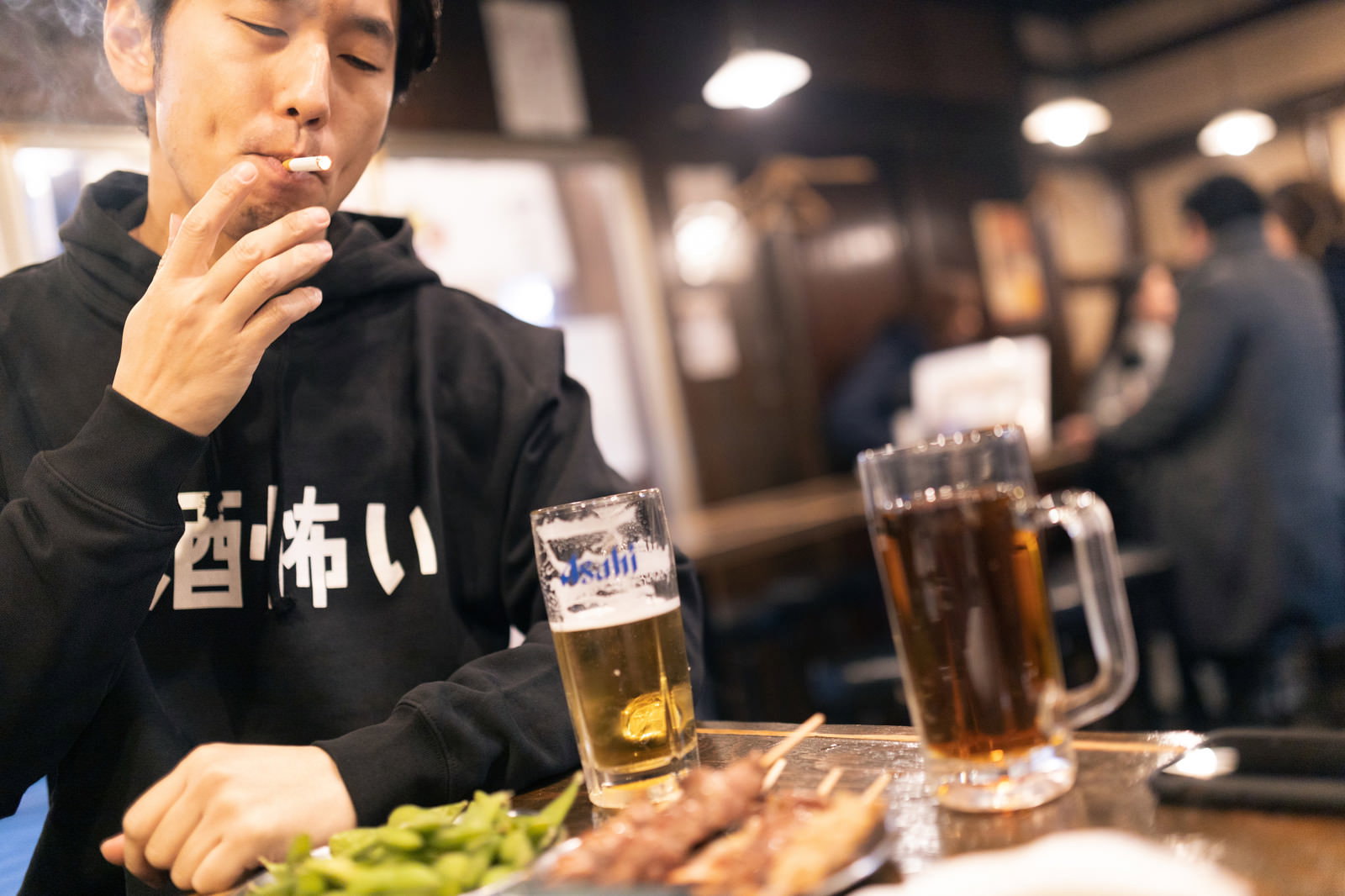 居酒屋のテーブルでタバコを吸う黒いパーカーの男性［モデル：大川竜弥］