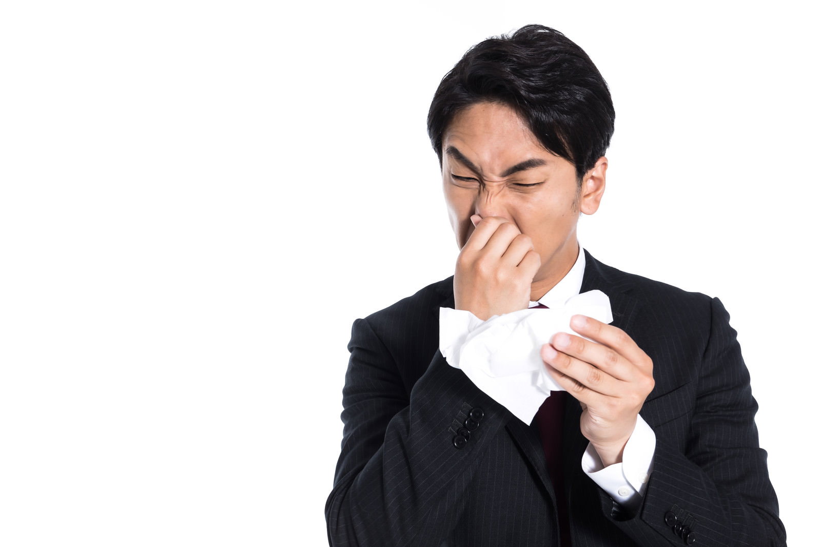 白背景でティッシュを鼻に当てて鼻をかんでいる黒いスーツ姿の男性［モデル：大川竜弥］
