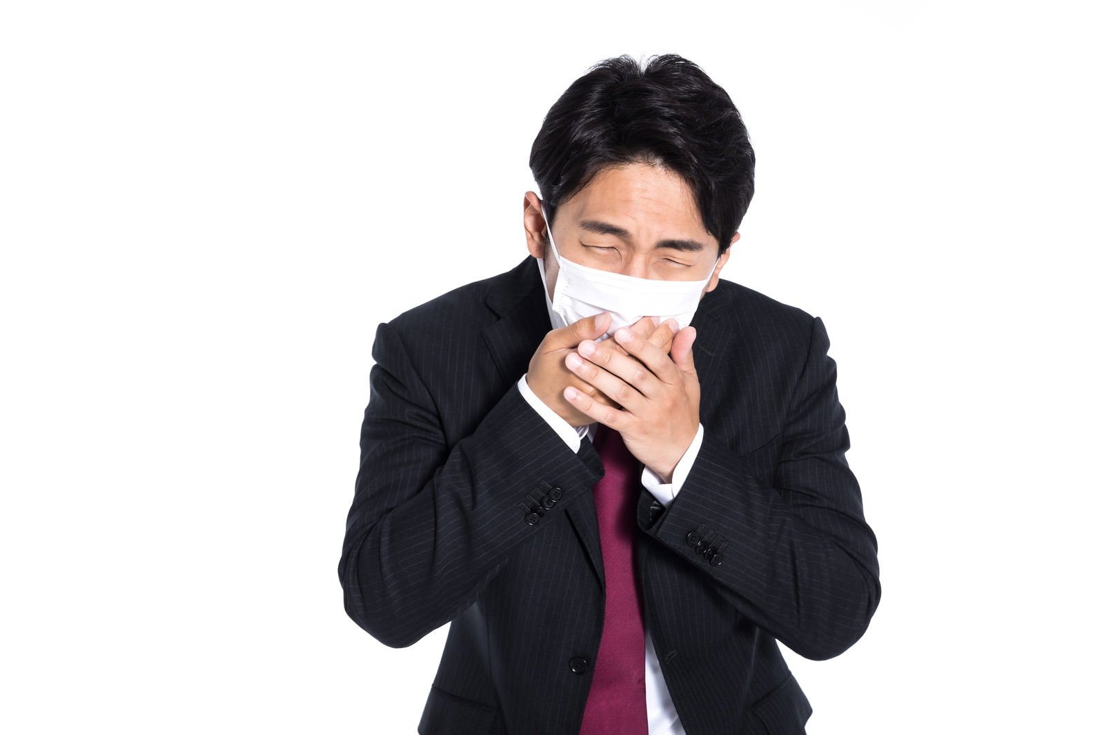 白いマスクを両手で押さえながら咳を我慢する黒いスーツの会社員男性［モデル：大川竜弥］