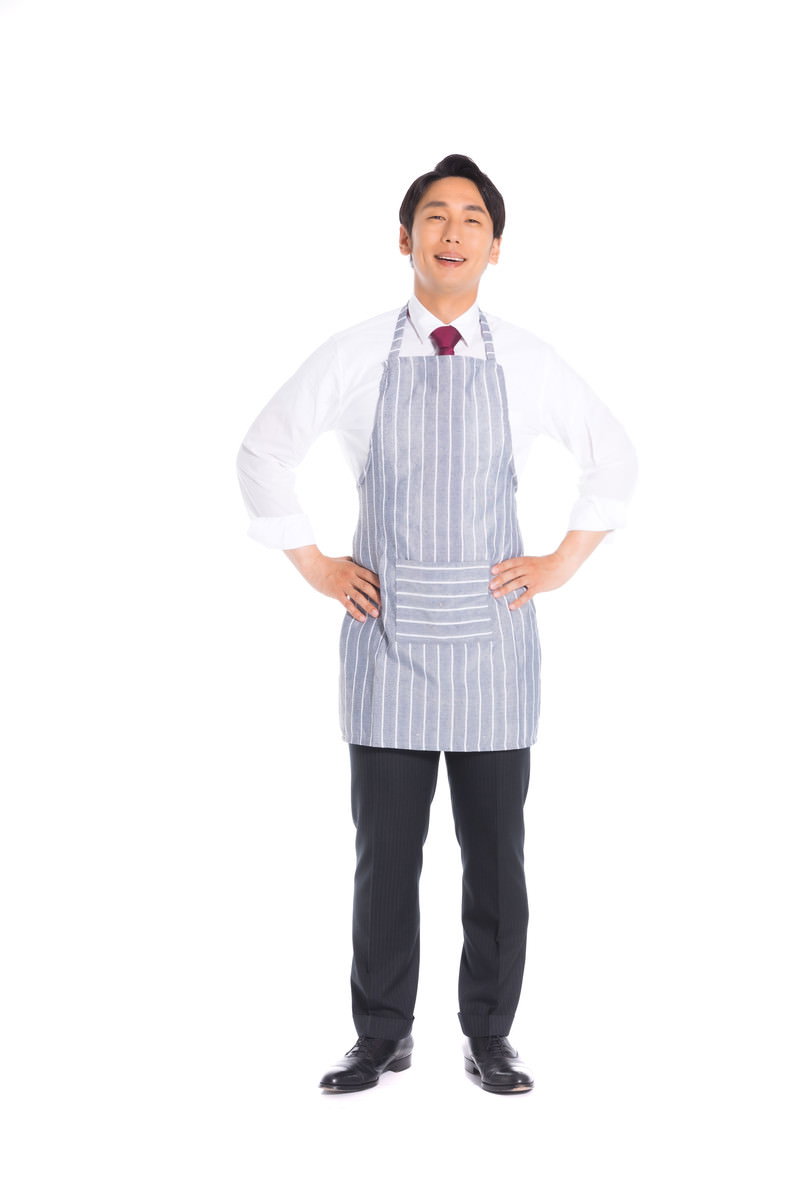 グレーのストライプエプロンを着用し腰に手を当てポーズをとる男性会社員［モデル：大川竜弥］