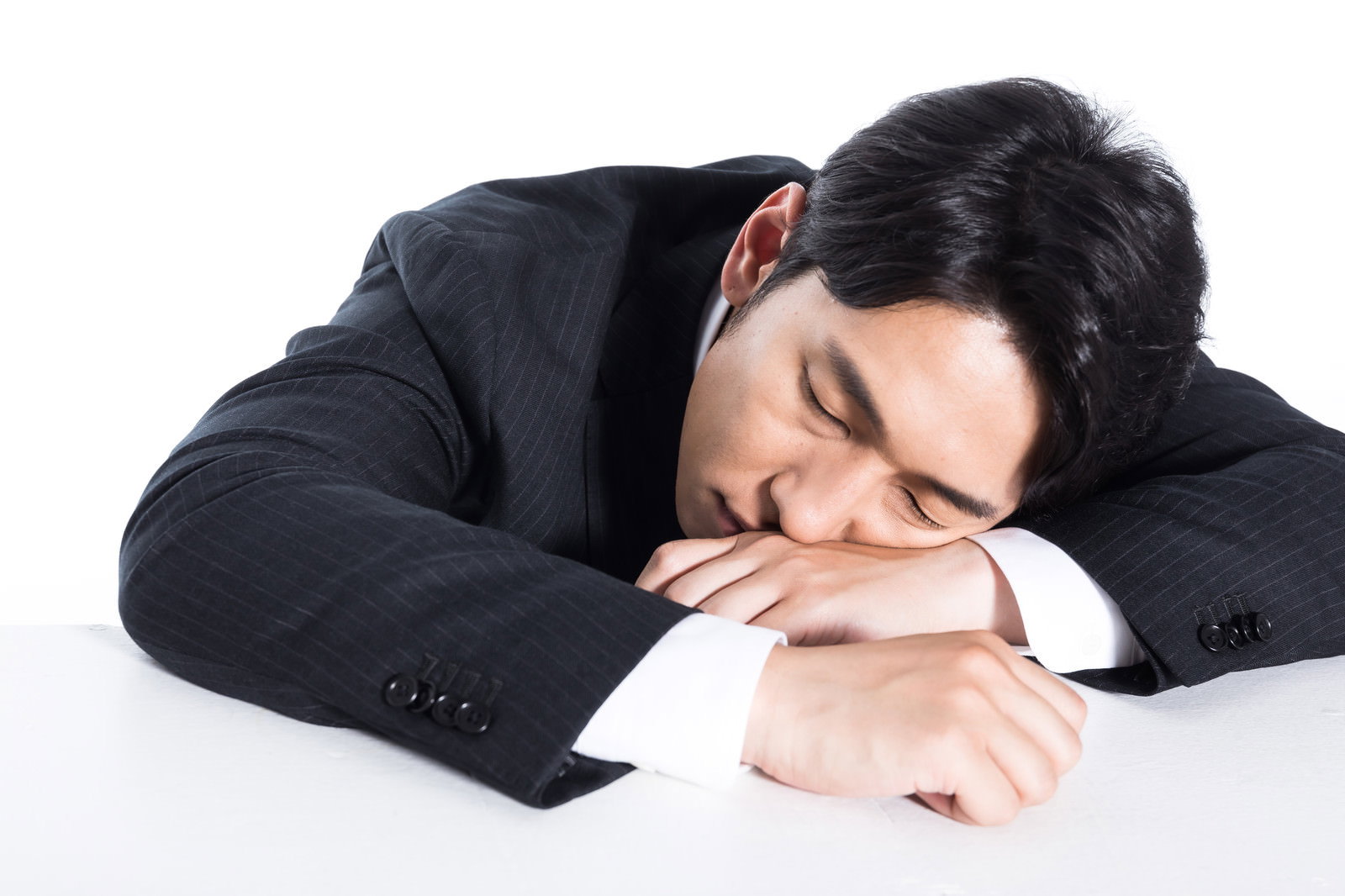 黒いスーツを着た男性が白い机の上で腕を枕にして居眠りしている［モデル：大川竜弥］