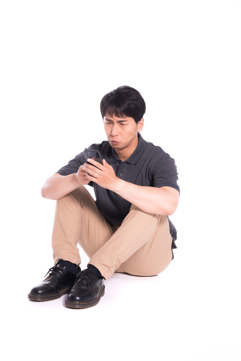 白い背景の前で体育座りをしながらスマートフォンを見る男性［モデル：大川竜弥］
