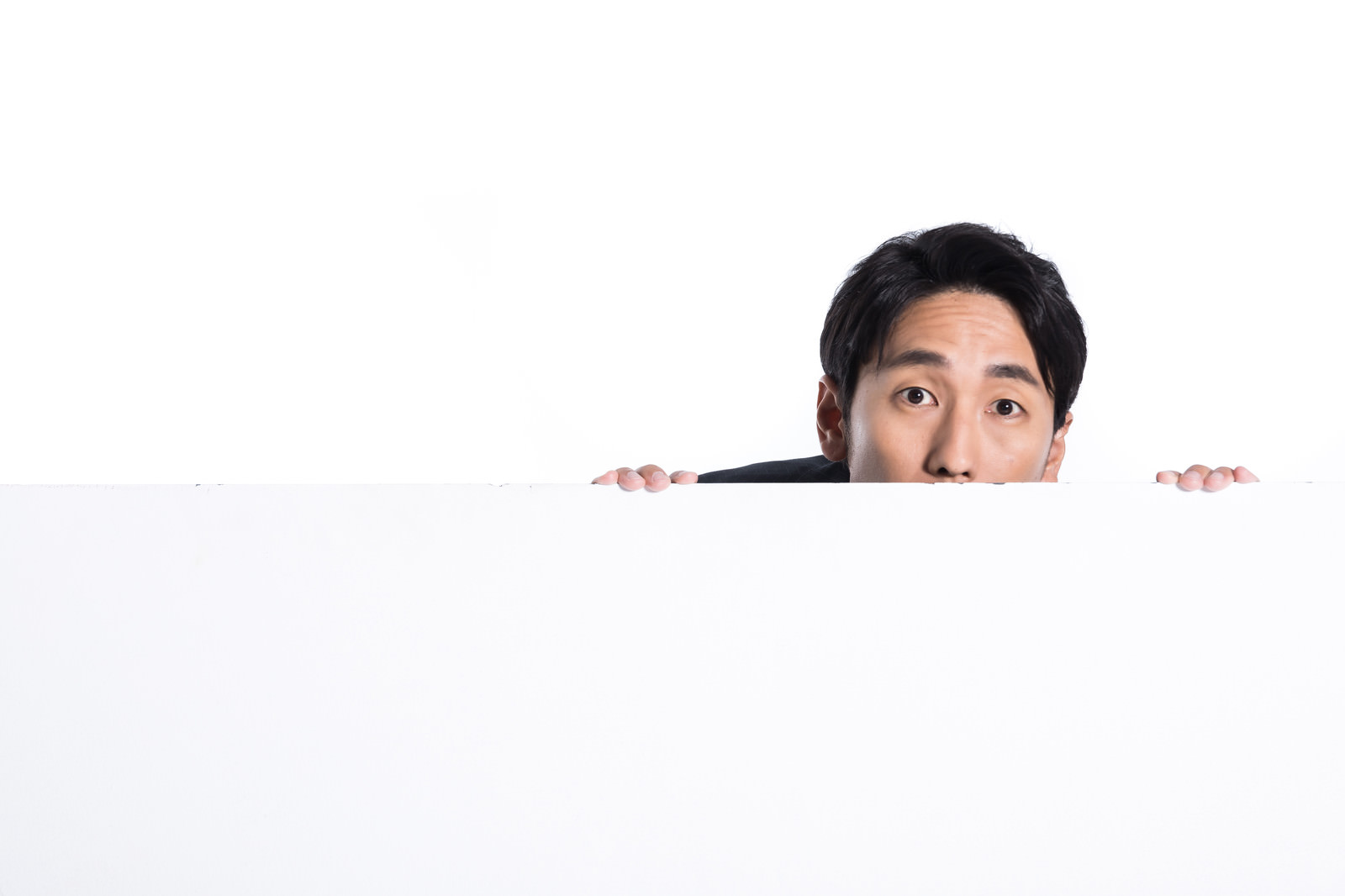白いホワイトボードの向こうから顔を出してのぞき見する男性［モデル：大川竜弥］
