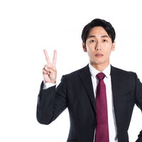 真顔でピースサインを見せるスーツ姿の男性の写真