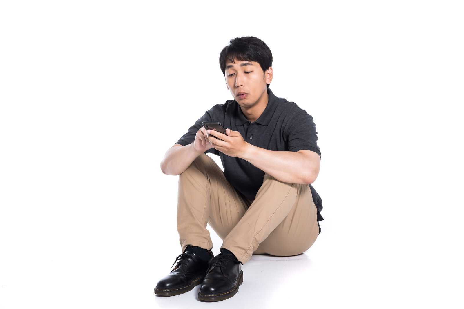 白い背景で体育座りしながらスマートフォンを見る男性［モデル：大川竜弥］