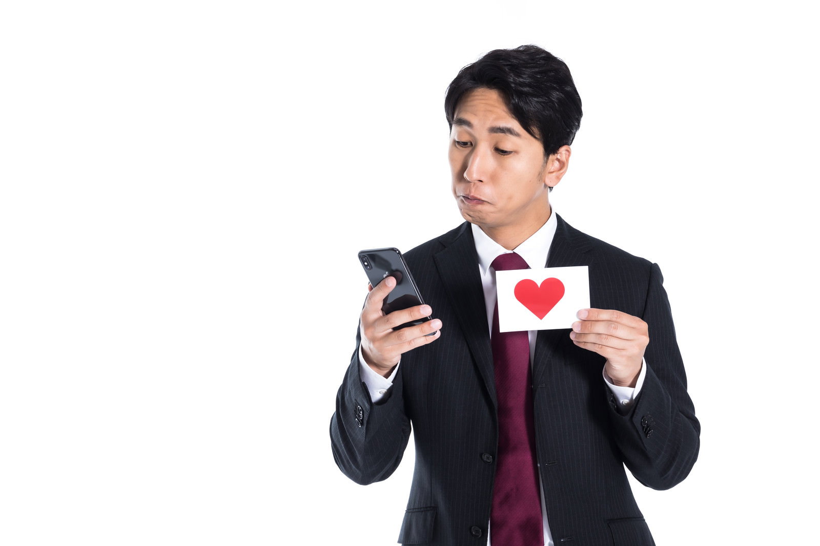 スマートフォンを見ながら赤いハートマークを持つスーツ姿の男性［モデル：大川竜弥］