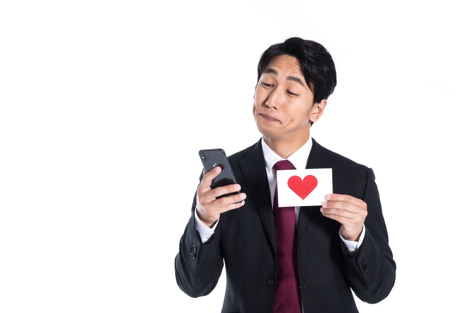 スマートフォンを見つめながらハートマークを持つスーツ姿の男性［モデル：大川竜弥］