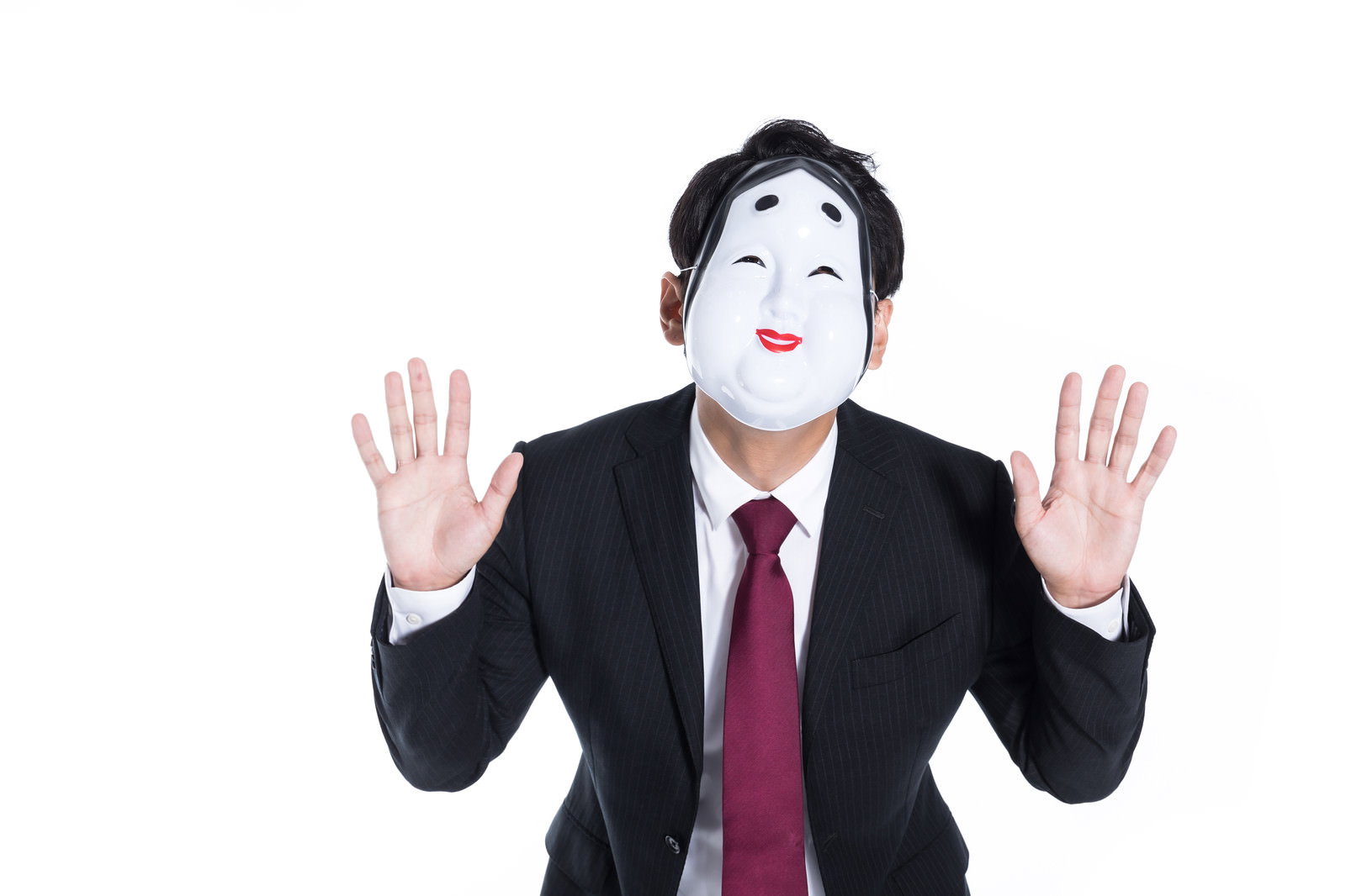 おかめのお面をつけて両手を上げるスーツ姿の男性［モデル：大川竜弥］