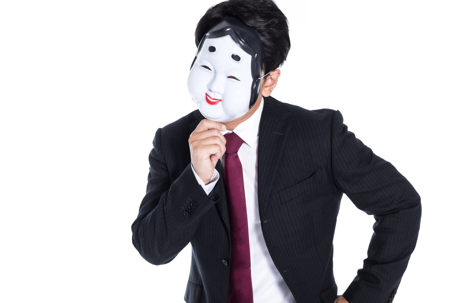 おかめの面を掲げて考え込むポーズをとるスーツ姿の男性［モデル：大川竜弥］