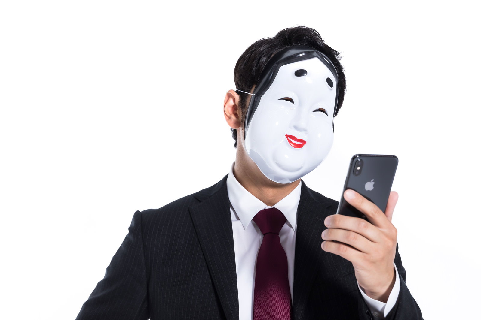 白い面をつけてスマートフォンを操作するスーツ姿の男性［モデル：大川竜弥］