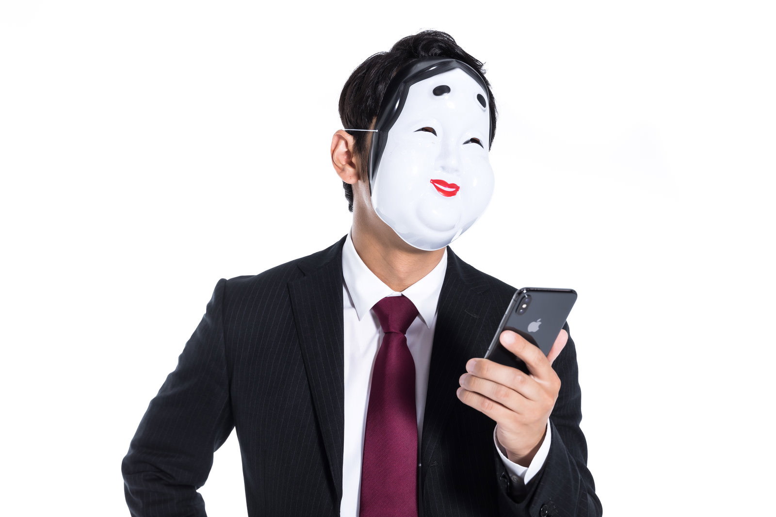 おかめのお面をつけてスマートフォンを持つスーツ姿の男性［モデル：大川竜弥］