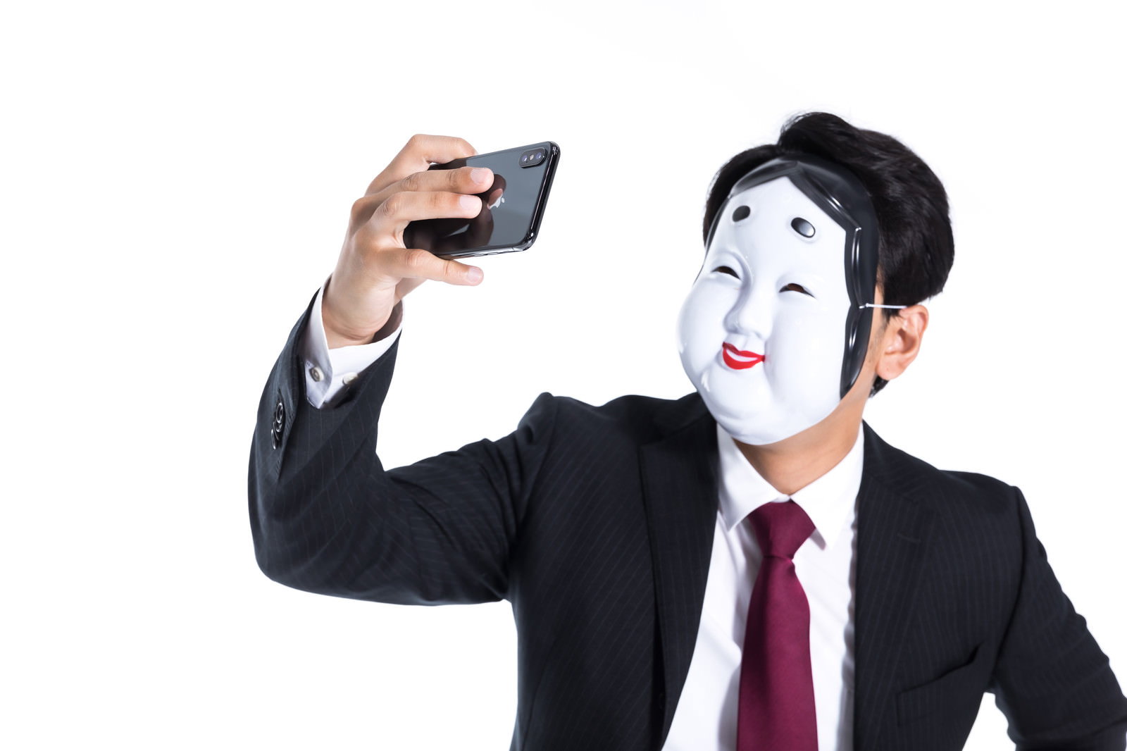 おかめのお面を被りスマートフォンで自撮りをする黒いスーツの男性［モデル：大川竜弥］