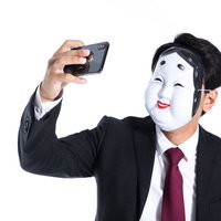 おかめのお面を着けてスマホで自撮りする詐欺男性の写真
