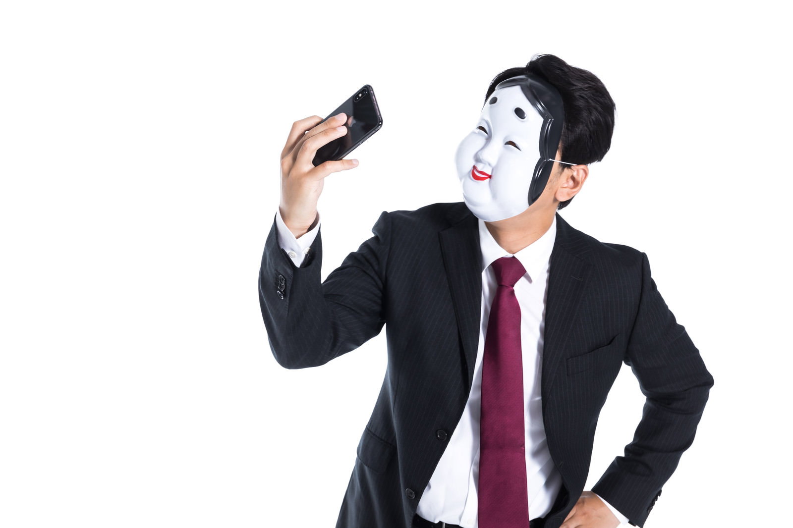 おかめの面をつけた黒いスーツの男性がスマートフォンで自撮りをしている［モデル：大川竜弥］