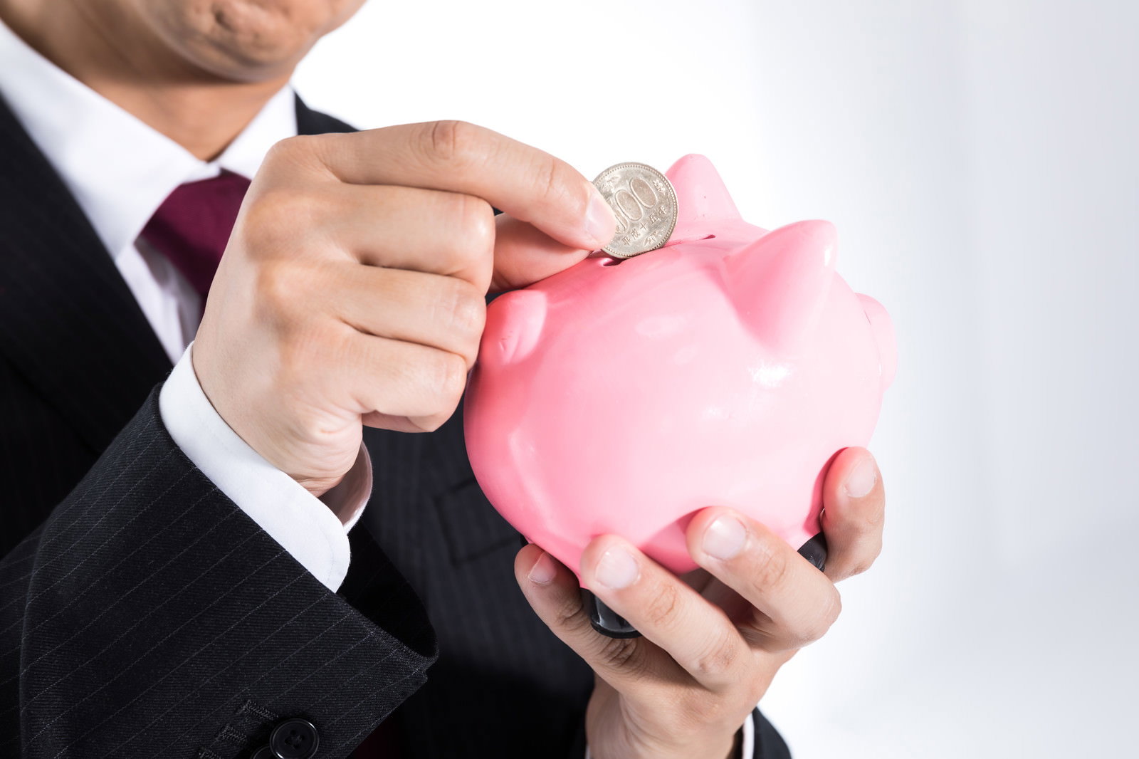 ピンク色の豚の貯金箱に硬貨を入れるスーツを着た男性の手元［モデル：大川竜弥］