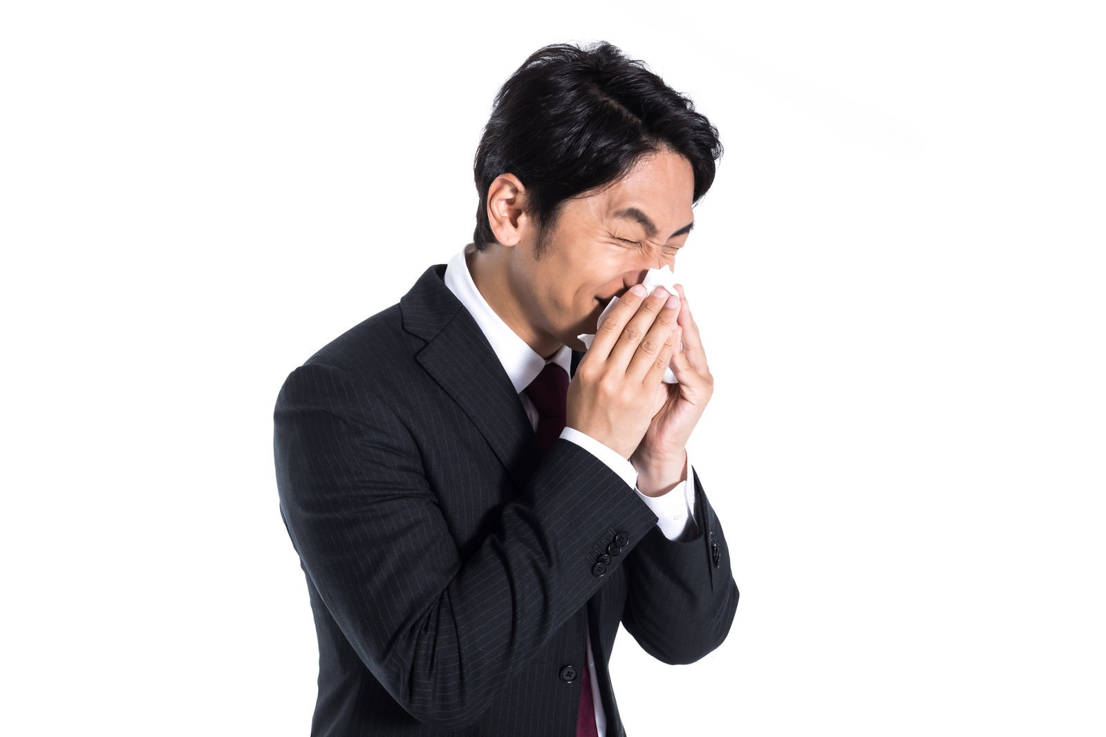 白い背景でティッシュを鼻に当てて鼻をかんでいる黒いスーツ姿の男性［モデル：大川竜弥］