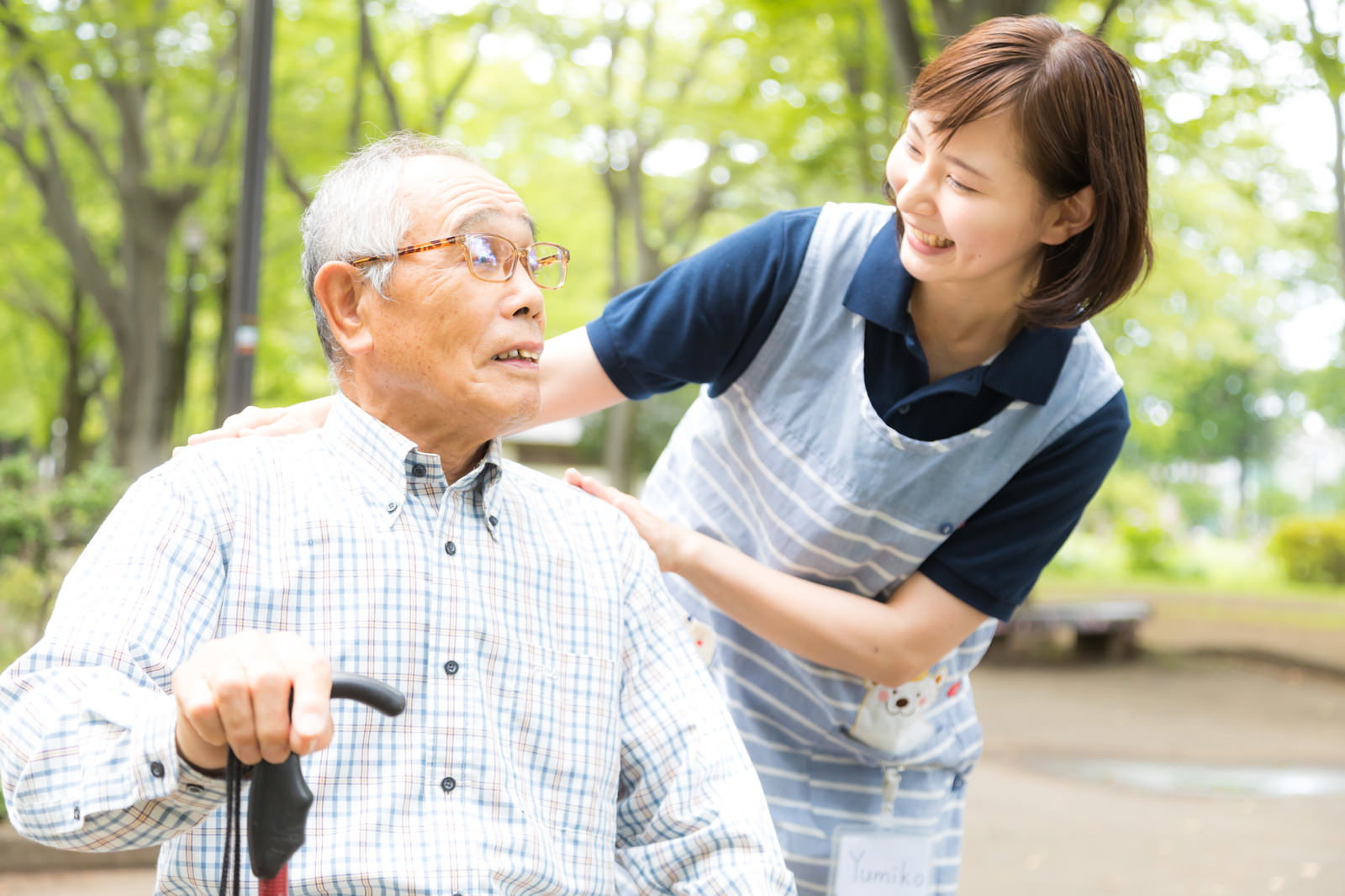 公園のベンチに座る高齢男性に優しく声をかける女性介護士［モデル：yumiko まーちゃん］