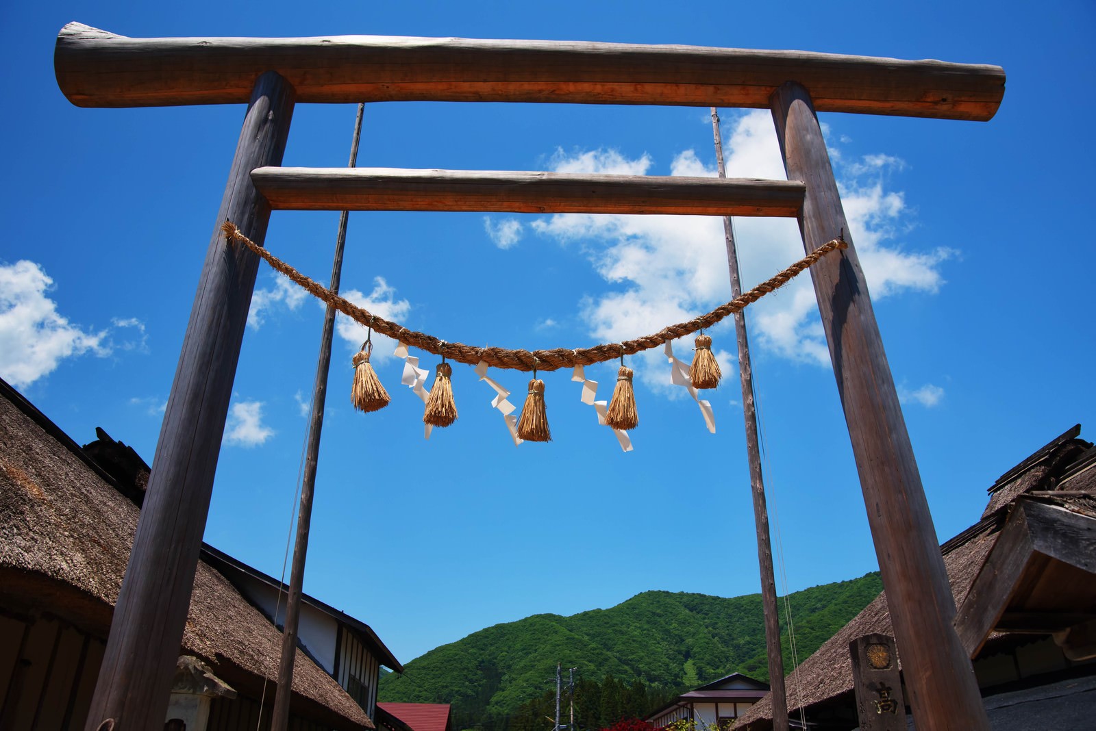 A wooden torii gate standing on the road lined with thatched-roof buildings in Ouchi-juku under the blue sky