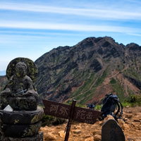 阿弥陀岳の山頂に立つ石造の標識と石像、連峰の景観の写真