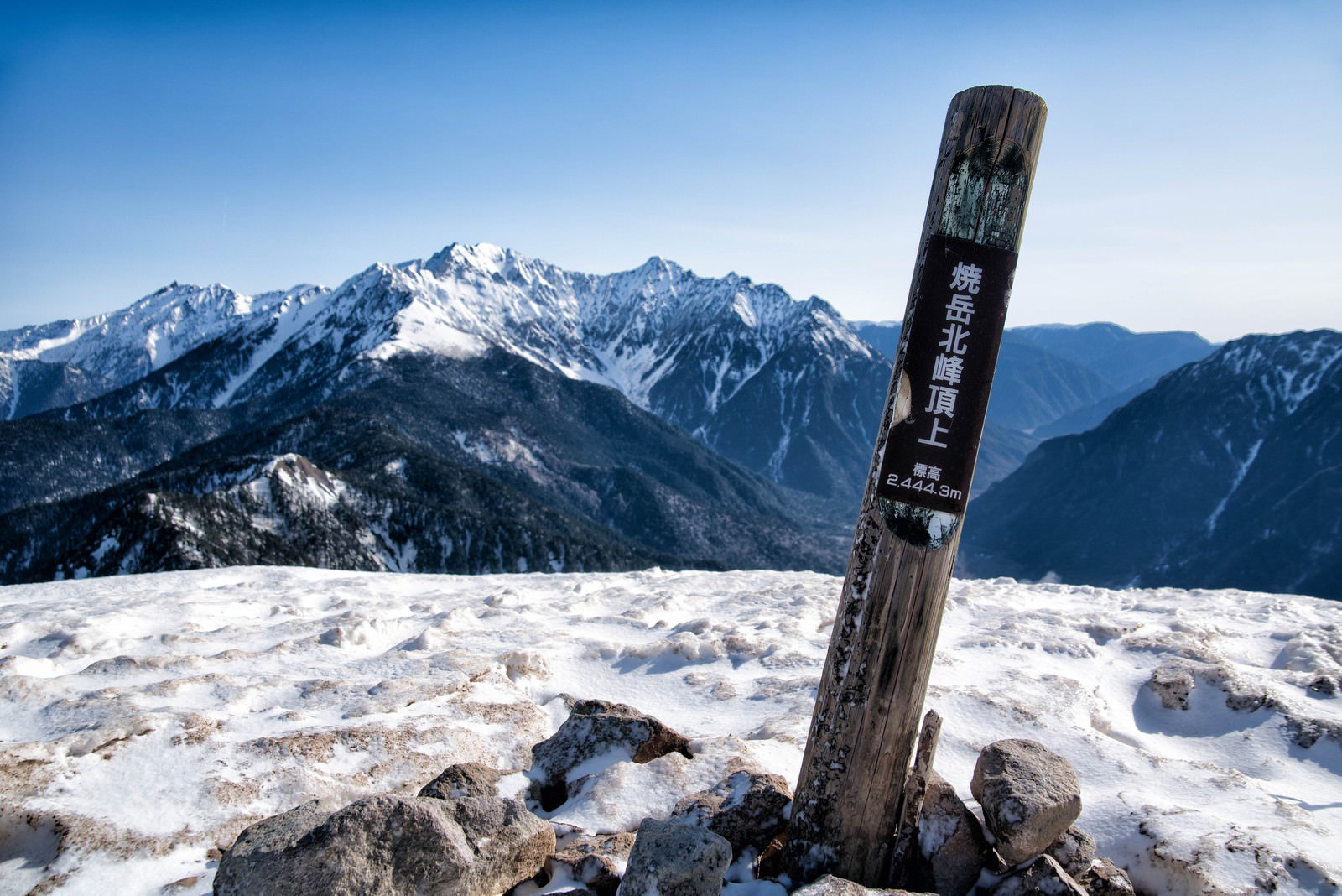 雪に覆われた焼岳山頂に立つ黒い標識と背景の北アルプスの山々
