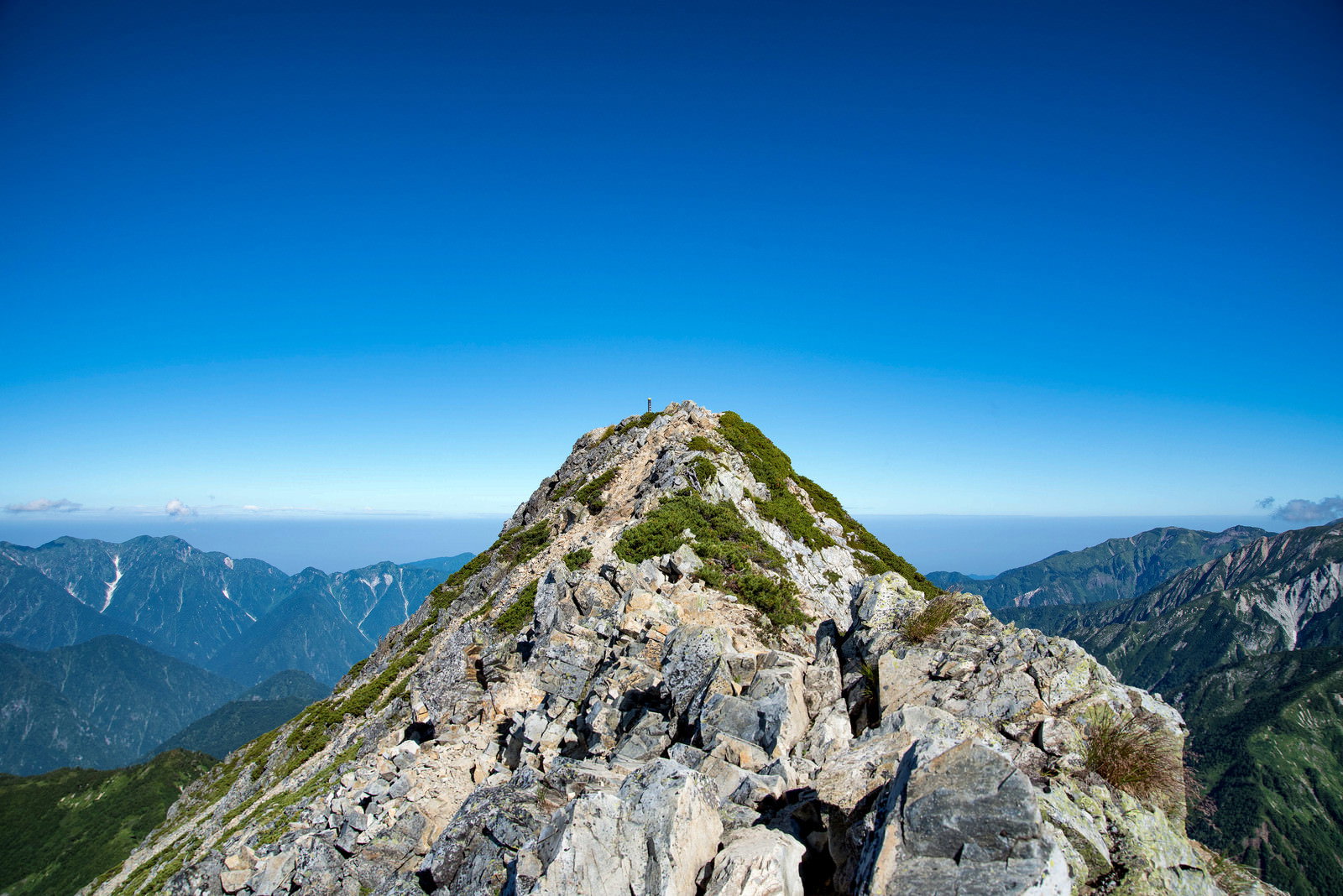 青空の下に切り立つ険しい岩峰の山頂と遠くの山脈