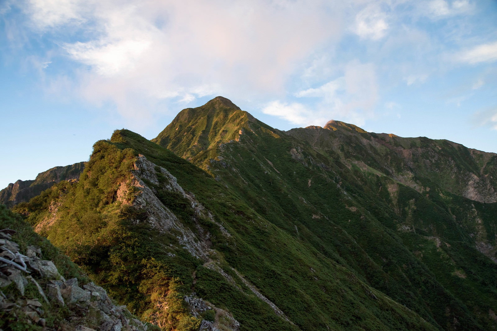 朝日を浴びる緑豊かな山の尾根と岩稜が青空の下に広がる山岳風景