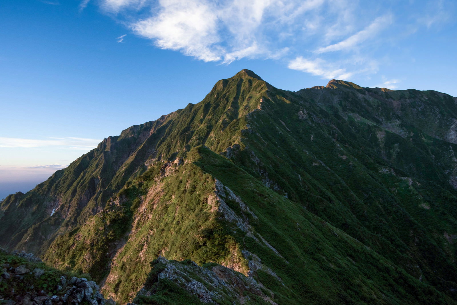 緑に覆われた険しい山稜線が連なり、青空に白い雲が浮かぶ山岳風景