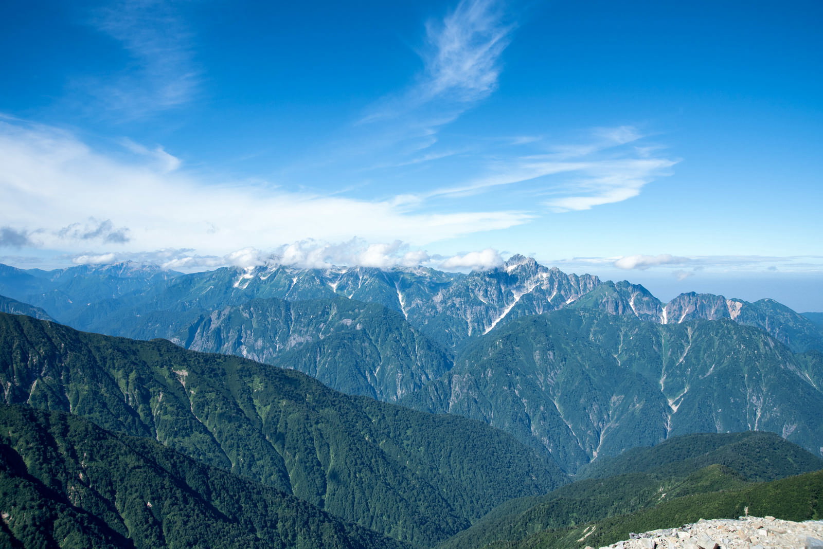 青空に白い雲が流れる中、険しい山々が連なる山岳連峰の風景