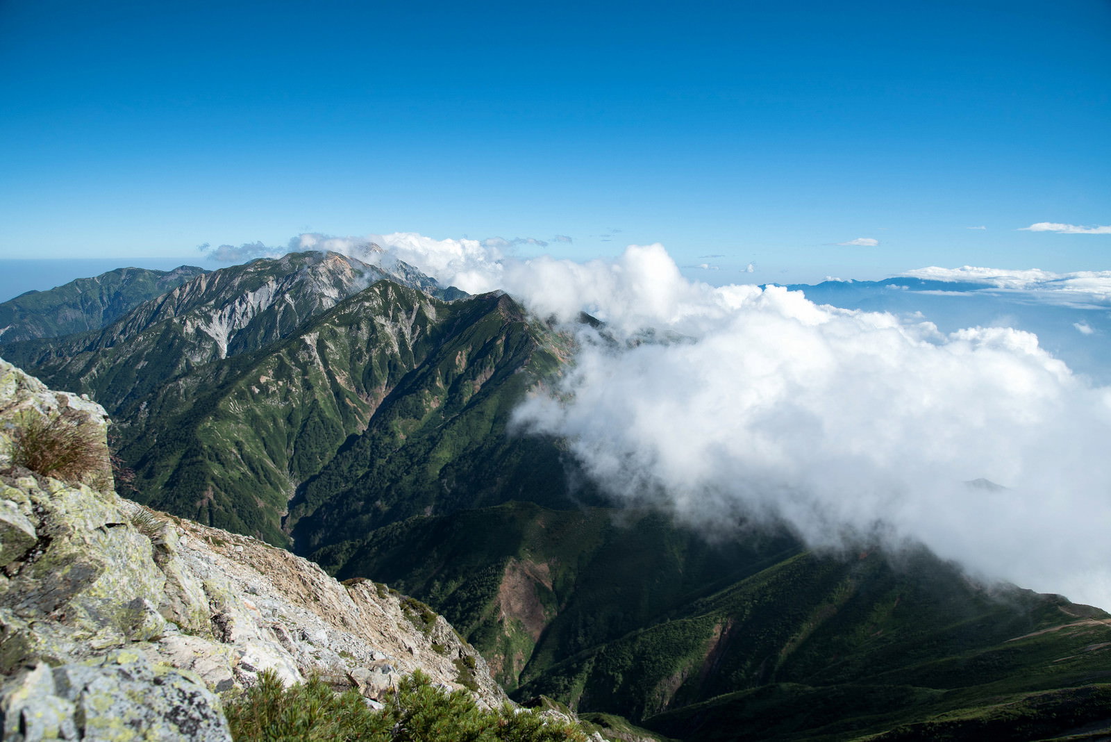 青空の下、緑の山稜に白い雲が流れ込む稜線の風景
