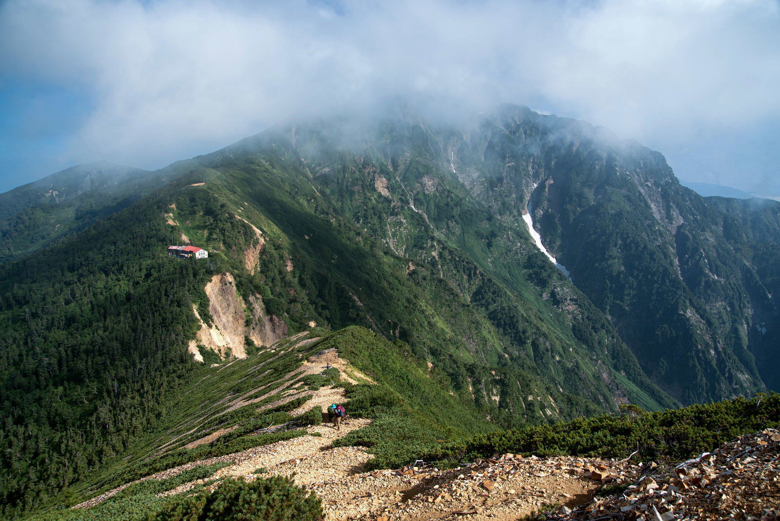 雲に包まれた険しい山々を背景に、山荘から登山道を歩く登山者