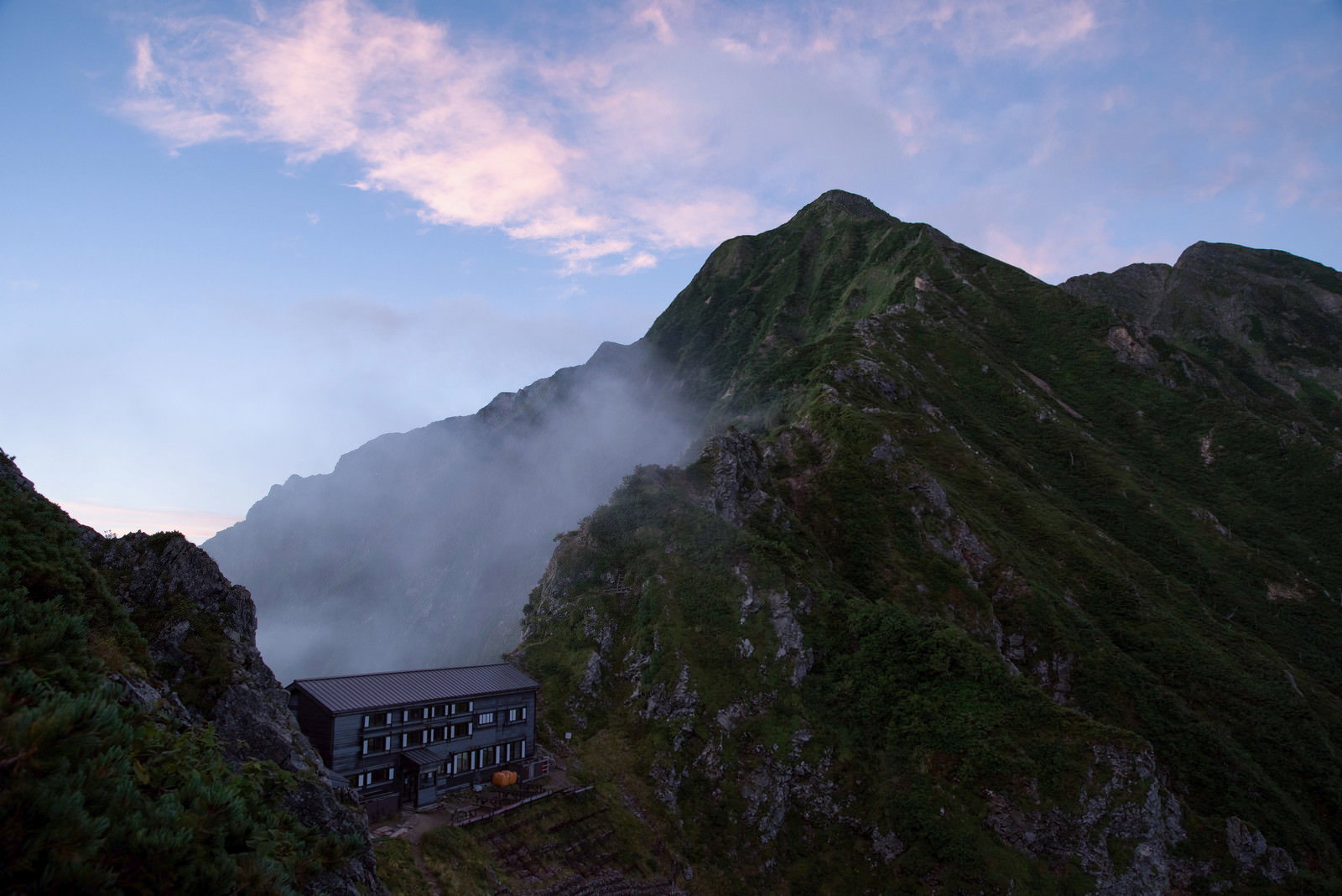 夕焼け空の下、険しい稜線に建つ山荘と登山道が見える山岳風景