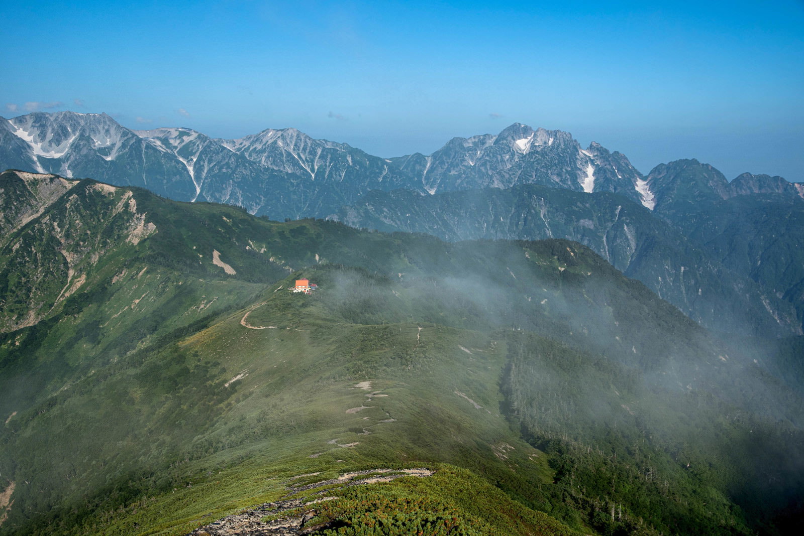 青空の下、緑の稜線を縫って続く登山道の先に山荘が見え、背景に連峰が広がる
