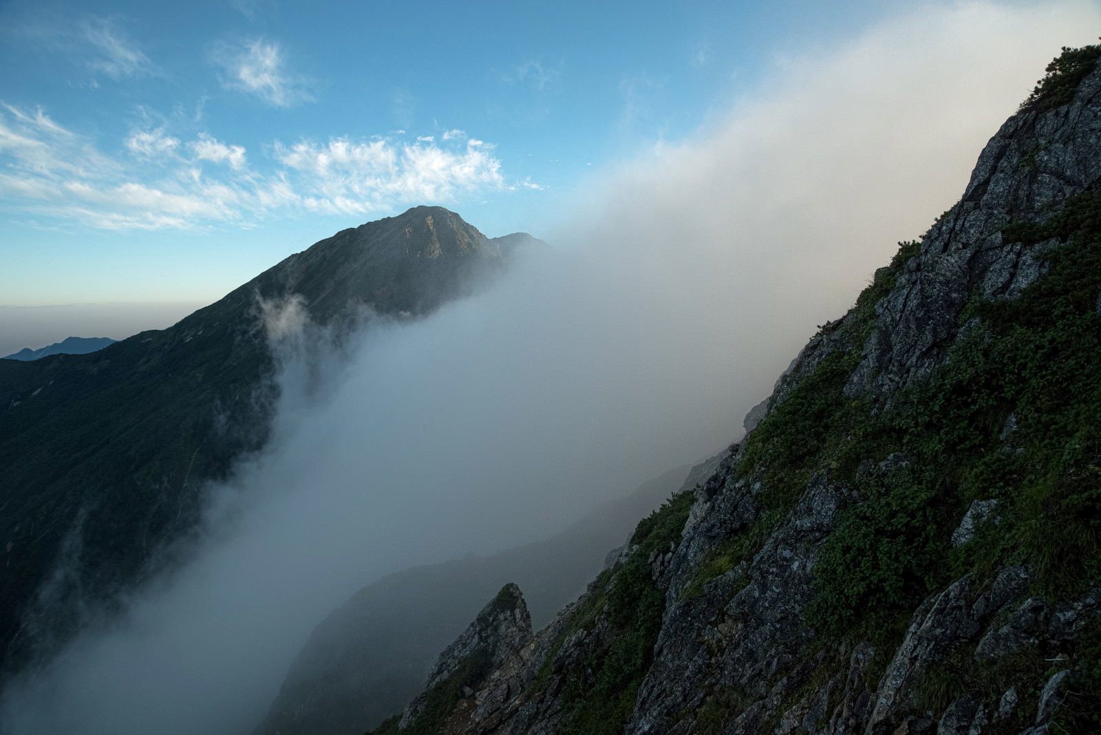 険しい岩尾根の向こうに白い雲が谷間へと流れ込む山岳風景
