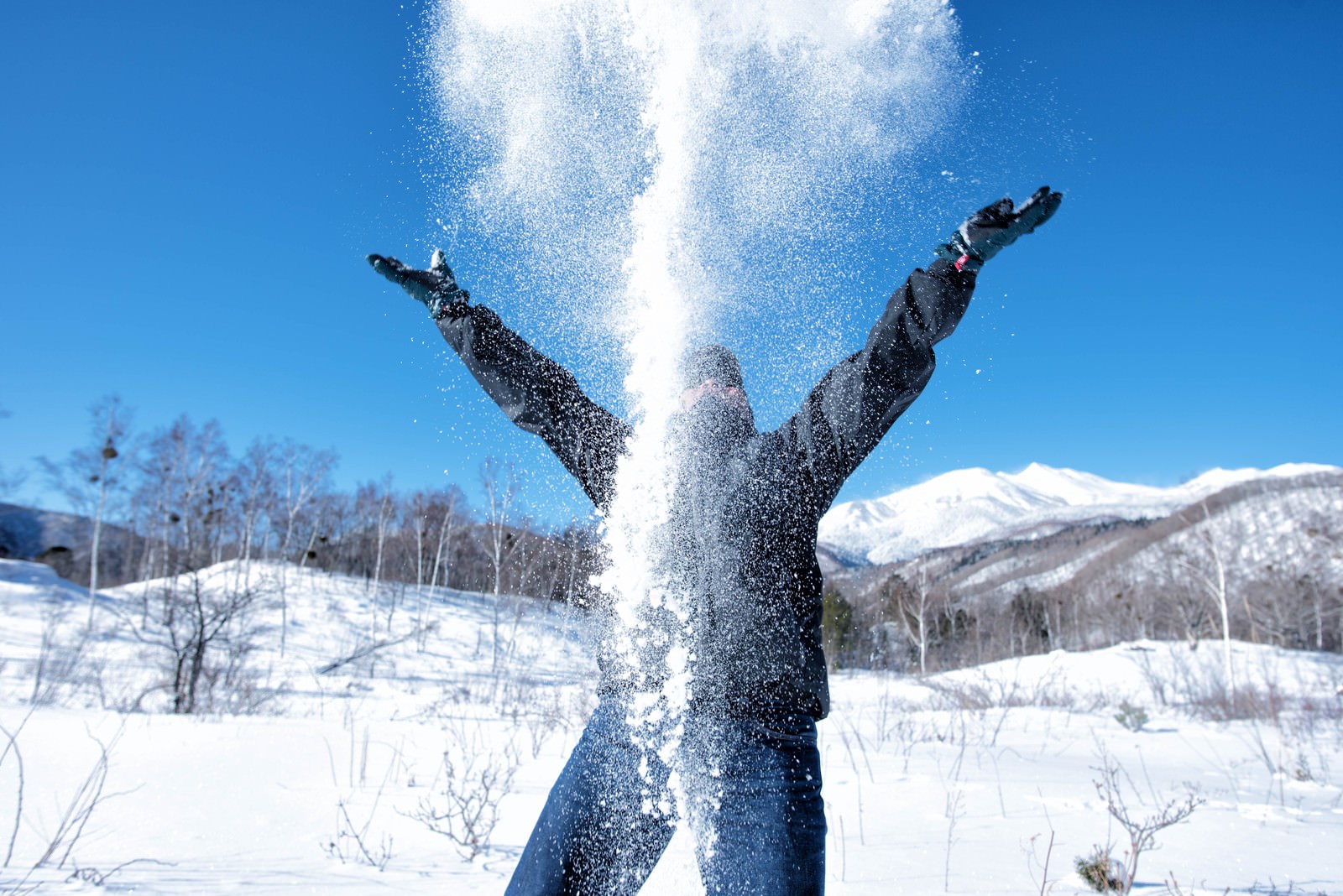 青空の下で両手を広げて雪を空に舞い上げるスキーウェア姿の人物