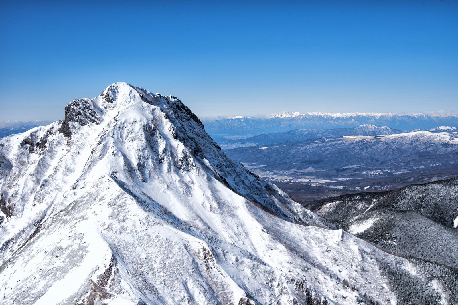 雪に覆われた阿弥陀岳の山頂と北アルプスの山脈が青空の下に広がる風景