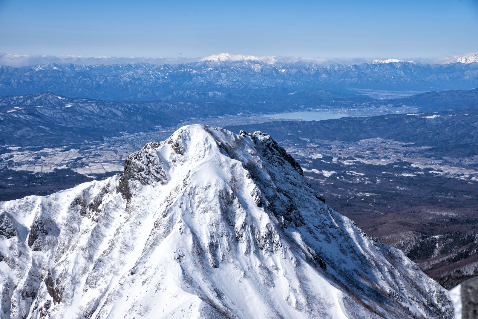 雪に覆われた阿弥陀岳の山頂から見える諏訪湖と青空の風景